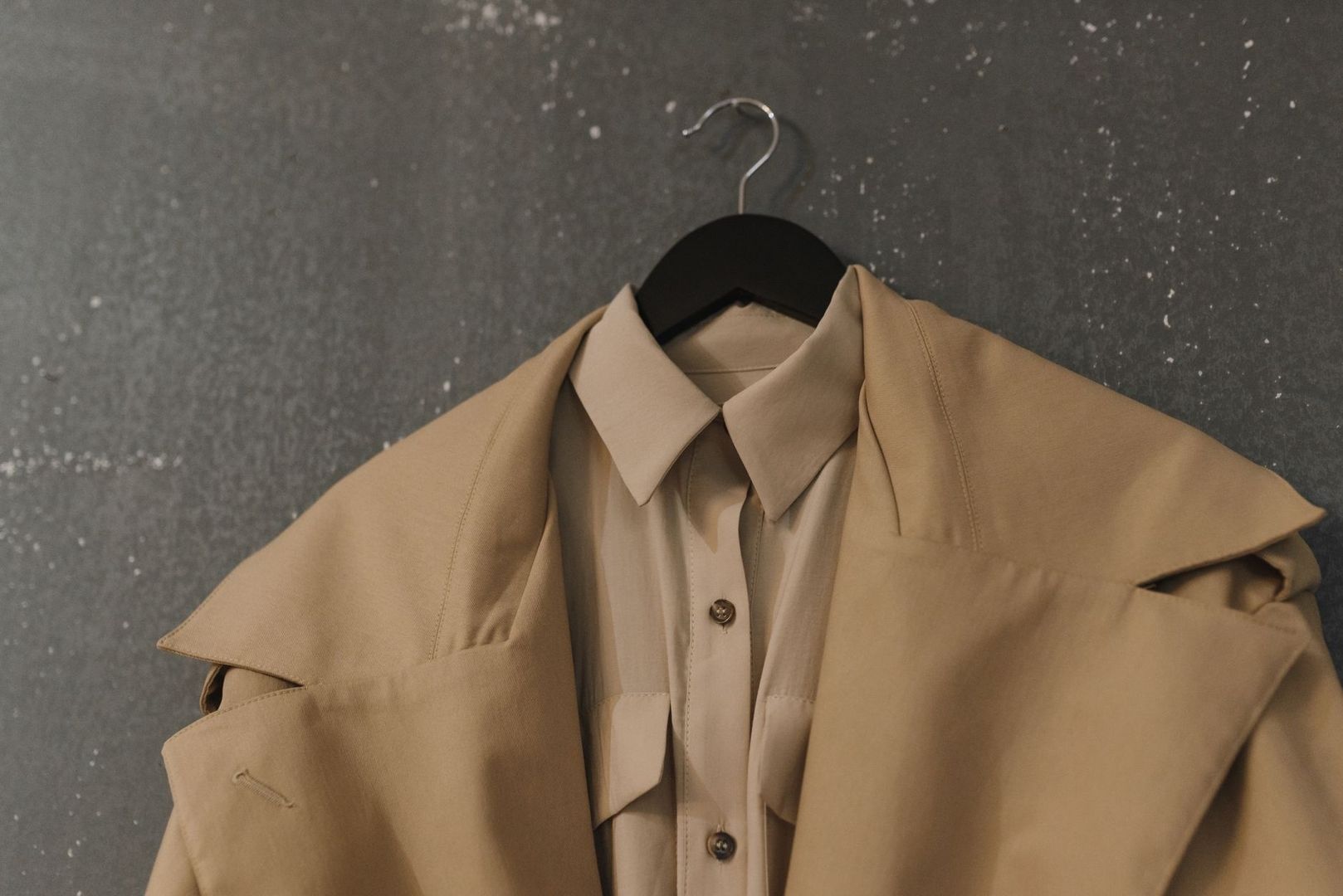 Beige trenchcoat och skjorta hänger på en svart galge mot en grå vägg